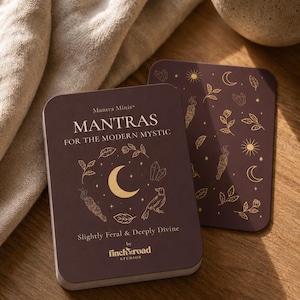 May include: Two rectangular cards with rounded corners featuring a dark purple background and gold foil illustrations of moons, stars, crystals, and birds. The text reads Mantra Minis, MANTRAS FOR THE MODERN MYSTIC, Slightly Feral & Deeply Divine, by finch road STUDIOS.