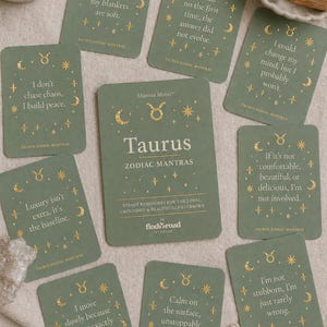 Könnte beinhalten: Eine Sammlung grüner Taurus-Zodiac-Mantra-Karten mit goldfarbenen Akzenten. Jede Karte zeigt ein Mantra und Himmelsillustrationen. Die zentrale Karte trägt die Aufschrift "Taurus ZODIAC MANTRAS". Die Karten sind auf einer neutralen Oberfläche angeordnet.