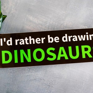 May include: A black rectangular sticker with the text "I'd rather be drawing DINOSAURS" in white and green. The sticker is on a gray surface with a green plant in the upper left corner.