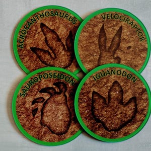 May include: Four round coasters with green edges and brown centers. Each coaster features a different dinosaur footprint and the dinosaur's name in green text. The dinosaurs are Acrocanthosaurus, Velociraptor, Sauroposeidon, and Iguanodon.