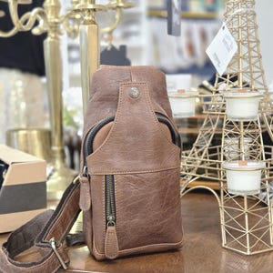 May include: A brown leather sling bag featuring a textured surface, a vertical zippered front pocket, a top zippered compartment, and a single adjustable shoulder strap. The bag is accented with dark metal hardware and a small circular metal stud.