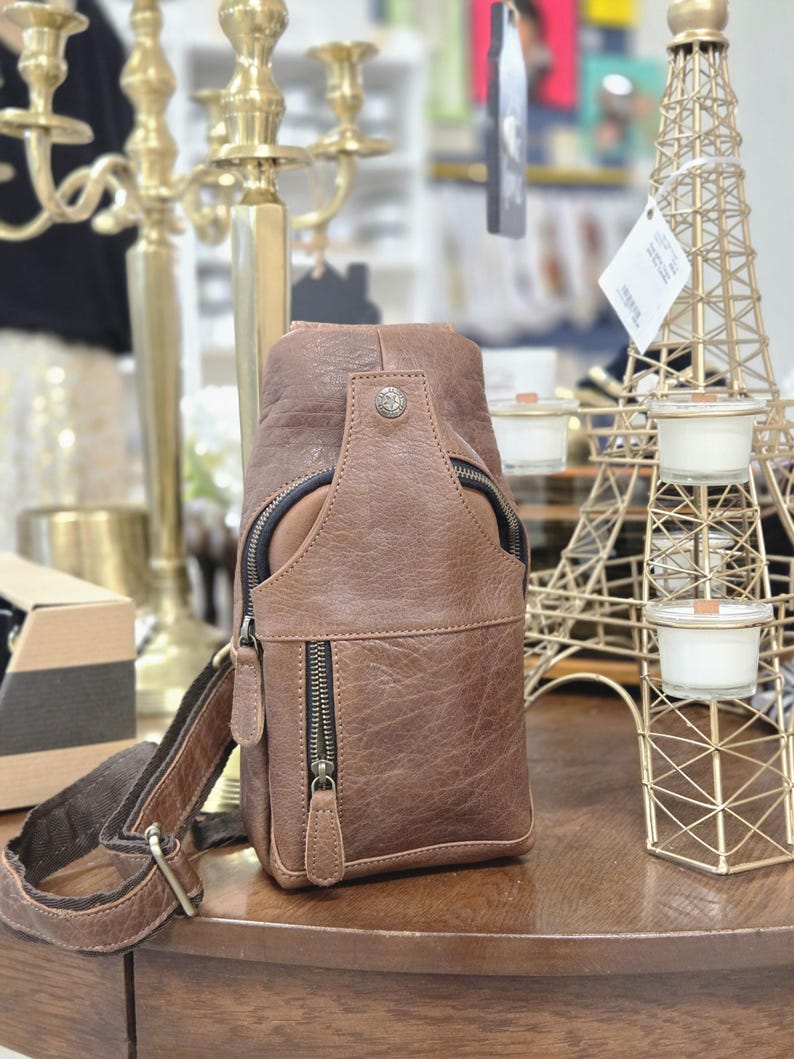 May include: A brown leather sling bag featuring a textured surface, a vertical zippered front pocket, a top zippered compartment, and a single adjustable shoulder strap. The bag is accented with dark metal hardware and a small circular metal stud.