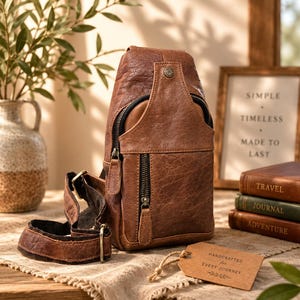 May include: A brown leather sling bag featuring a vertical zippered front pocket, a top flap with a metal snap button, and an adjustable strap. A rectangular cardboard tag is attached, printed with the text HANDCRAFTED for EVERY JOURNEY.