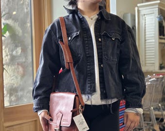 May include: A brown leather messenger bag with a flap closure and adjustable shoulder strap. The bag has a buckle closure and a tag hanging from the strap. The bag is worn over a black denim jacket.