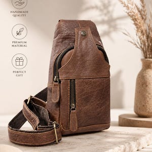 May include: A brown textured leather sling bag featuring a vertical zippered front pocket, a main zippered compartment, and an adjustable strap with metal hardware. The bag includes a small circular metal stud detail near the top closure.