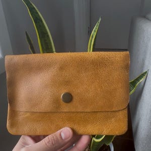 Peut inclure: Une pochette rectangulaire en cuir jaune moutarde avec une surface texturée et un bouton-pression central de couleur laiton. La pochette présente un design à rabat avec des coins arrondis et une finition lisse et uniforme sur le devant.