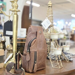 May include: A brown leather sling bag featuring a vertical zippered front pocket, a main compartment with a dark zipper, and an adjustable brown fabric strap with metal hardware. The bag has a textured finish and a small metal stud detail at the top.