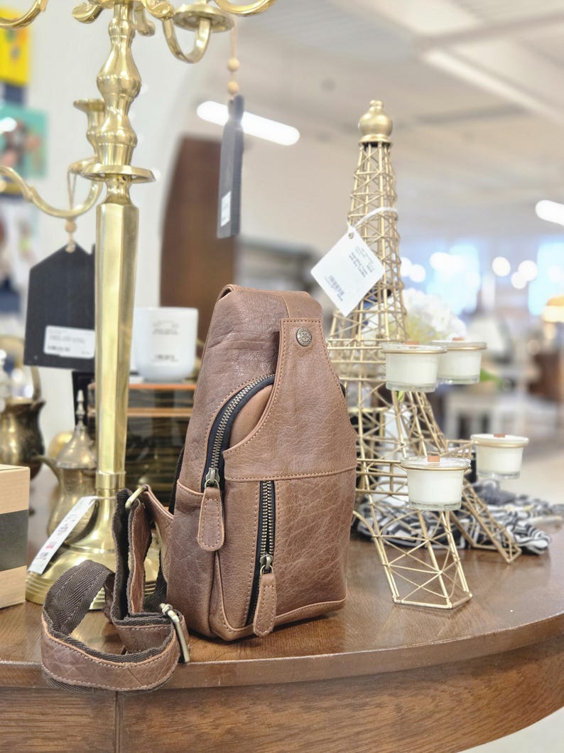 May include: A brown leather sling bag featuring a vertical zippered front pocket, a main compartment with a dark zipper, and an adjustable brown fabric strap with metal hardware. The bag has a textured finish and a small metal stud detail at the top.