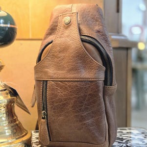 May include: A brown textured leather sling bag featuring a vertical zippered front pocket, a top flap with a metallic snap button closure, and dark metal hardware. The bag has a structured, rounded silhouette with visible stitching along the edges.