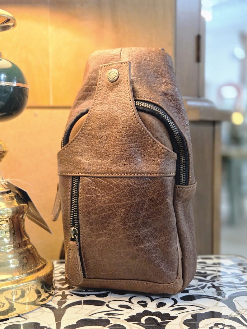 May include: A brown textured leather sling bag featuring a vertical zippered front pocket, a top flap with a metallic snap button closure, and dark metal hardware. The bag has a structured, rounded silhouette with visible stitching along the edges.