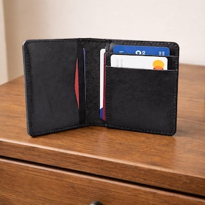 Puede incluir: Cartera de cuero negro abierta con múltiples ranuras para tarjetas. La cartera está abierta, revelando un interior rojo y varias tarjetas de crédito. La cartera está sobre una superficie de madera.