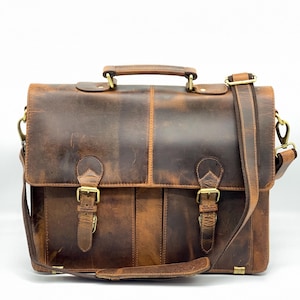 May include: A brown leather briefcase with a handle and shoulder strap. The bag features two front buckles and brass-coloured hardware. The leather has a distressed appearance.