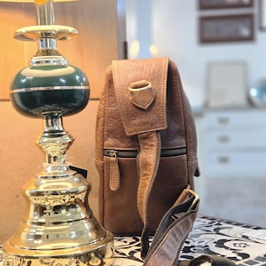 May include: A brown leather sling bag with a textured surface, a front zipper pocket, and an adjustable fabric strap with metal hardware, positioned next to a lamp with a green and gold base and a light-colored lampshade.