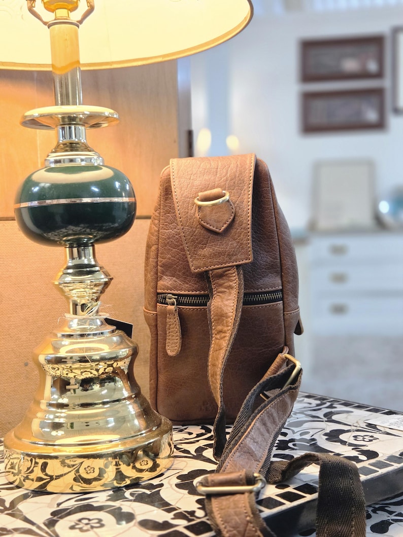 May include: A brown leather sling bag with a textured surface, a front zipper pocket, and an adjustable fabric strap with metal hardware, positioned next to a lamp with a green and gold base and a light-colored lampshade.
