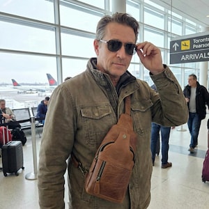 May include: A tan leather sling bag with a front zippered pocket and a metal snap closure, worn across the chest over a brown utility jacket. The bag features a single adjustable shoulder strap and visible stitching details on the textured leather surface.