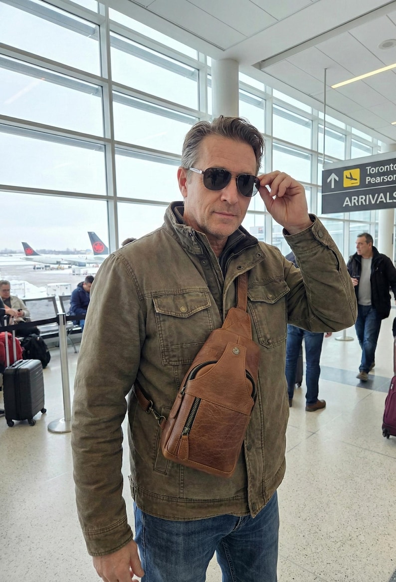 May include: A tan leather sling bag with a front zippered pocket and a metal snap closure, worn across the chest over a brown utility jacket. The bag features a single adjustable shoulder strap and visible stitching details on the textured leather surface.