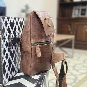 May include: A brown leather sling bag featuring a textured surface, a front zippered pocket with a matching leather pull tab, a top flap with a brass-toned D-ring attachment, and an adjustable fabric strap connected to the side of the bag.