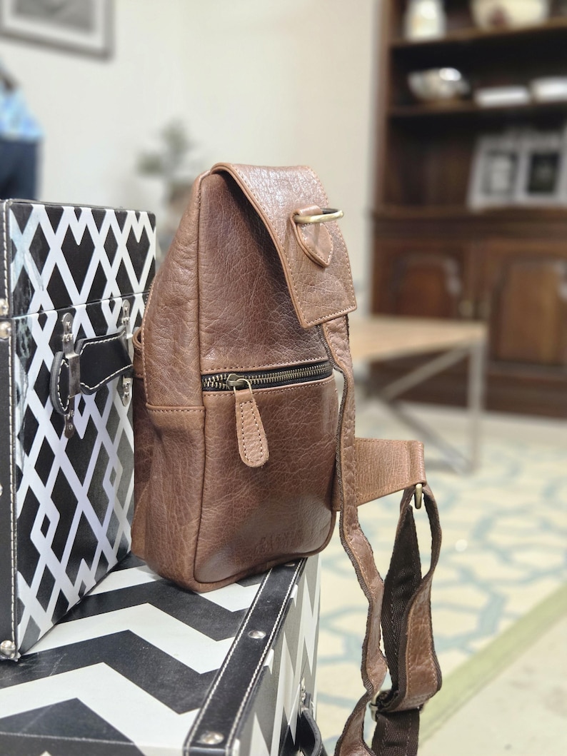 May include: A brown leather sling bag featuring a textured surface, a front zippered pocket with a matching leather pull tab, a top flap with a brass-toned D-ring attachment, and an adjustable fabric strap connected to the side of the bag.