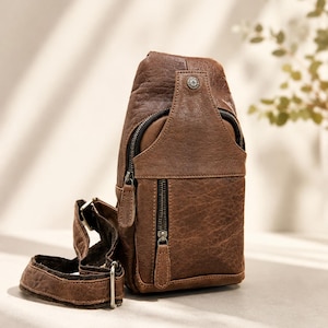 May include: A brown leather sling bag featuring a textured finish, a vertical zippered front pocket, a main zippered compartment, and an adjustable strap with metal hardware. A small circular metal emblem is attached to the top front section.