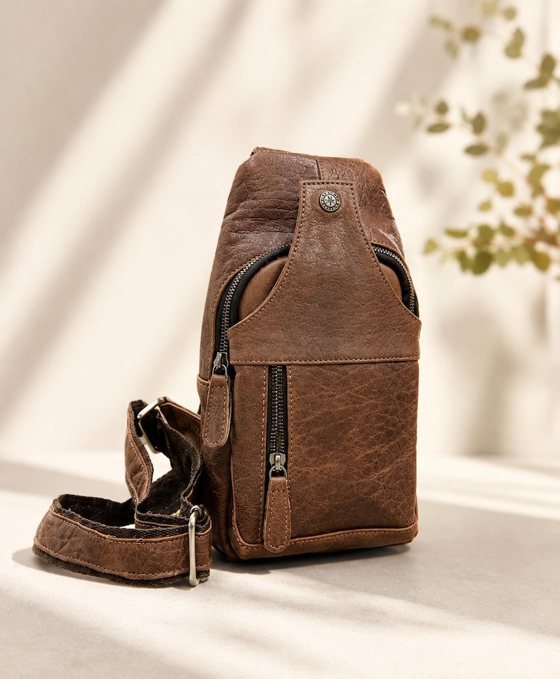 May include: A brown leather sling bag featuring a textured finish, a vertical zippered front pocket, a main zippered compartment, and an adjustable strap with metal hardware. A small circular metal emblem is attached to the top front section.