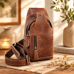 May include: A brown leather sling bag featuring a textured surface, multiple zippered compartments with black lining, a vertical front pocket, and an adjustable brown fabric strap with antique brass-toned metal hardware and a small circular stud.
