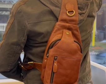 Puede incluir: Un bolso bandolera de cuero marrón con un bolsillo frontal con cremallera y un bolsillo más pequeño con cremallera. El bolso tiene una correa para el hombro con un anillo de metal color latón. El bolso se lleva cruzado.