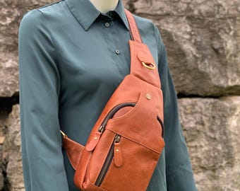 Puede incluir: Un bolso bandolera de cuero marrón con múltiples compartimentos con cremallera y una correa para el hombro. El bolso se lleva cruzado sobre el pecho y presenta un anillo metálico dorado y tiradores de cremallera. El bolso se muestra en un maniquí con una camisa verde azulado.