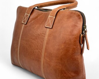 May include: A brown leather tote bag with a top handle and a zip closure. The bag has white stitching and a rectangular shape. The leather has a textured appearance. The bag is designed for carrying items.