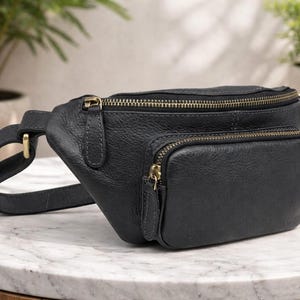May include: A black leather bum bag with two zipped compartments and a smaller front pocket. The bag features gold-coloured zips and hardware. The adjustable strap is attached with a gold-coloured buckle. The bag is on a marble surface.