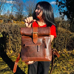 May include: A dark brown leather backpack with a roll-top closure, a central vertical strap featuring a brass-toned metal buckle, a horizontal front pocket flap, and a side vertical zippered pocket with a leather pull tab and adjustable straps.