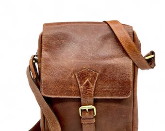 May include: A brown leather messenger bag with a flap closure and adjustable shoulder strap. The bag features a buckle closure and brass hardware. The leather has a distressed appearance.