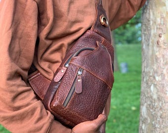 Puede incluir: Un bolso bandolera de cuero marrón con una superficie texturizada y múltiples compartimentos con cremallera. El bolso tiene una correa para el hombro y un anillo metálico de color latón. El bolso se lleva cruzado.