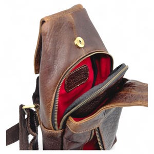 May include: A brown leather crossbody bag with a red interior. The bag has a zippered main compartment and an adjustable strap. The leather has a distressed look, and the bag is open, revealing the interior.
