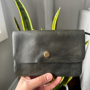 May include: A dark green leather wallet with a brass button closure. The wallet is rectangular and has a flap. The leather has a slightly distressed appearance. The wallet is held in front of a potted plant.