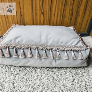 May include: A gray rectangular dog bed with a decorative trim and tassels around the edges.