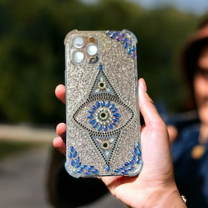 May include: A silver glitter phone case with a blue evil eye design. The case has a clear back and a raised edge around the camera lens.