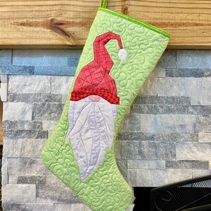 Peut inclure: Une chaussette de Noël matelassée, ornée d'un motif de gnome aux couleurs rouge, blanc et vert. Le gnome porte un bonnet rouge et une barbe blanche. La chaussette est suspendue à une cheminée en bois, prête pour les fêtes.
