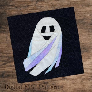Puede incluir: Un bloque de edredón de tela cuadrado con un diseño de fantasma creado con la técnica de costura sobre papel. El fantasma es blanco con sutiles acentos de estilo acuarela en morado y azul, ojos rectangulares negros y una boca negra.