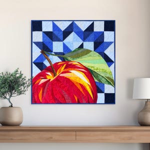 Könnte beinhalten: Ein quadratischer Wandbehang aus Stoff mit einem Patchwork-Design, das einen großen roten Apfel mit gelben und orangen Akzenten sowie ein grünes Blatt zeigt. Der Hintergrund besteht aus einem geometrischen Muster aus blauen Quadraten.