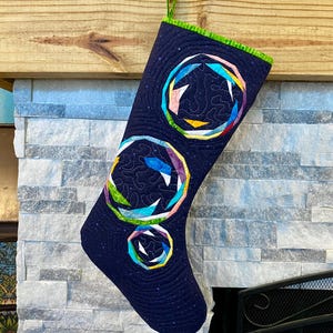 May include: A navy blue quilted Christmas stocking with three colourful geometric designs. The stocking has a green trim and is hanging from a wooden mantel. The geometric designs are made of various colours, including pink, yellow, blue, and green.