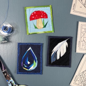 Puede incluir: Tres bloques de tela para acolchar con diseños de piezas: un hongo rojo y blanco sobre fondo azul claro, una gota de agua azul y verde sobre fondo azul oscuro y una pluma blanca sobre fondo negro, junto con patrones de papel.