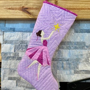 May include: A quilted Christmas stocking in shades of lavender and pink, featuring a ballerina design. The ballerina is reaching for a yellow star. The stocking is hanging from a wooden mantel.