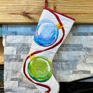 Może przedstawiać: Ręcznie robiona pończocha świąteczna z białym tłem, z dwoma kolorowymi, geometrycznymi wzorami ozdób. Górna ozdoba jest niebieska, a dolna zielona, z czerwonymi akcentami wstążki i czerwoną lamówką.