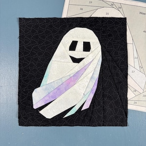 Könnte beinhalten: Ein quadratischer Stoff-Quiltblock mit einer Geister-Applikation aus weißen, hellvioletten und hellblauen Stoffstücken auf einem schwarzen Hintergrund mit dezentem Muster. Eine Papiervorlage für das Nähen ist hinter dem Block sichtbar.