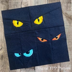 May include: A square fabric quilt block featuring a dark navy blue textured background with four sets of eyes pieced into the design. Two eyes are bright yellow, one set is orange with black pupils, and one set is light blue with black pupils.
