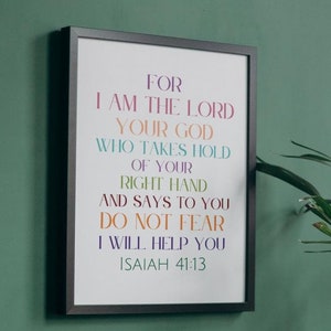Puede incluir: Una impresión enmarcada con un marco negro y un fondo blanco. La impresión presenta una cita colorida de la Biblia, Isaías 41:13, "Porque yo soy el Señor tu Dios, que te tomo de la mano derecha y te digo: No temas, yo te ayudaré." La impresión está en una pared verde con una planta en maceta delante. El texto debajo de la impresión dice "Combina con múltiples esquemas de colores."