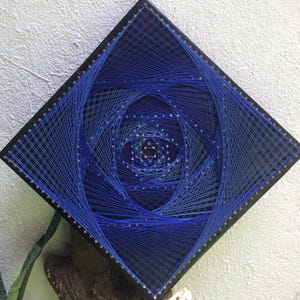 May include: A square string art piece with a black frame. The artwork features a blue spiral design created with blue thread on a black background. The piece is mounted on a white textured wall.