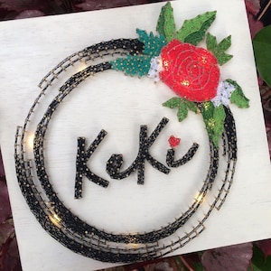 Può includere: Un'opera d'arte a filo fatta a mano su una tavola di legno bianca. Il design presenta un cerchio nero a pois con luci incorporate, una rosa rossa, foglie verdi e il nome "Keki" in lettere nere a pois. Un oggetto decorativo unico.