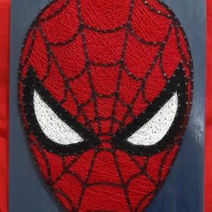 Può includere: Un'opera d'arte a filo fatta a mano raffigurante la maschera di Spider-Man. Il design presenta filo rosso che forma il volto della maschera, con occhi bianchi e contorni neri. L'opera è montata su una tavola di legno blu.