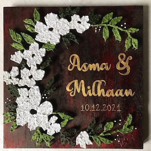 Può includere: Opera d'arte da parete quadrata in legno con sfondo rosso scuro. Fiori bianchi in filo e foglie verdi formano un bordo. Il testo dorato recita "Asma & Milhaan" e "10.12.2021".